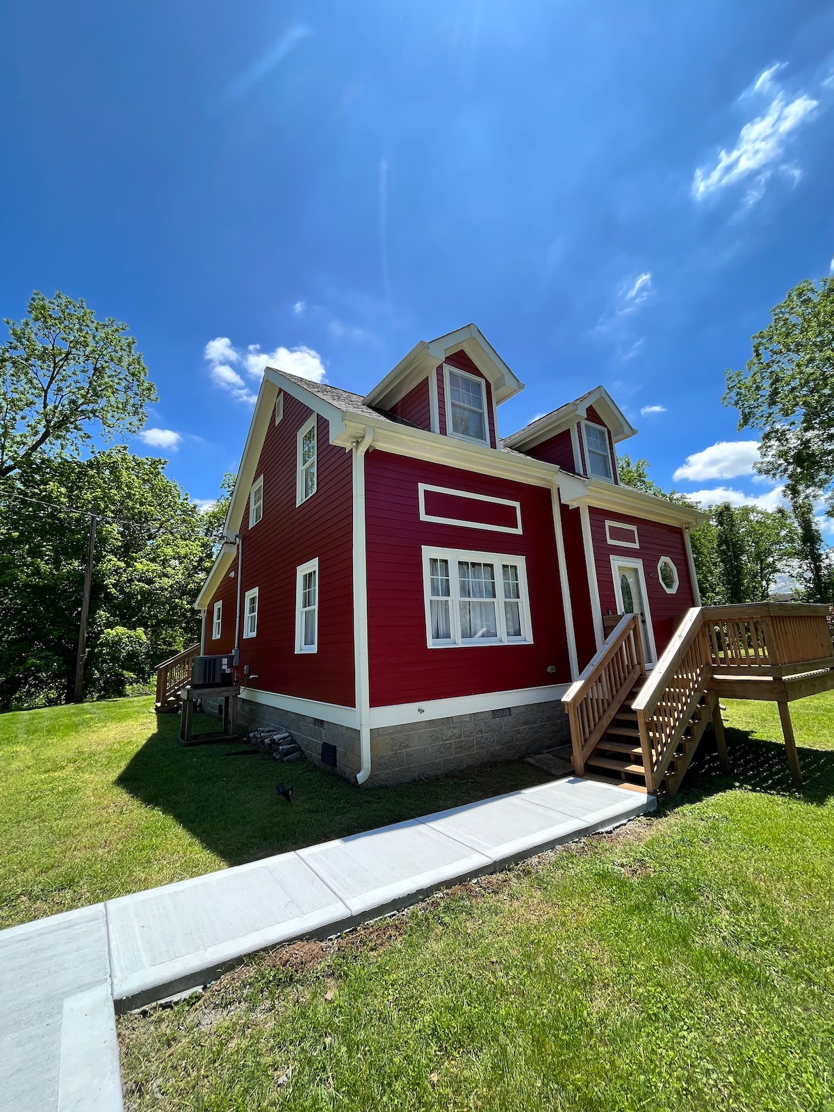 The exterior of a charming red house with a classic design is photographed under a clear blue sky. Lush green grass surrounds the property, while steps lead up to the entrance. A pathway runs alongside, providing access to the yard.