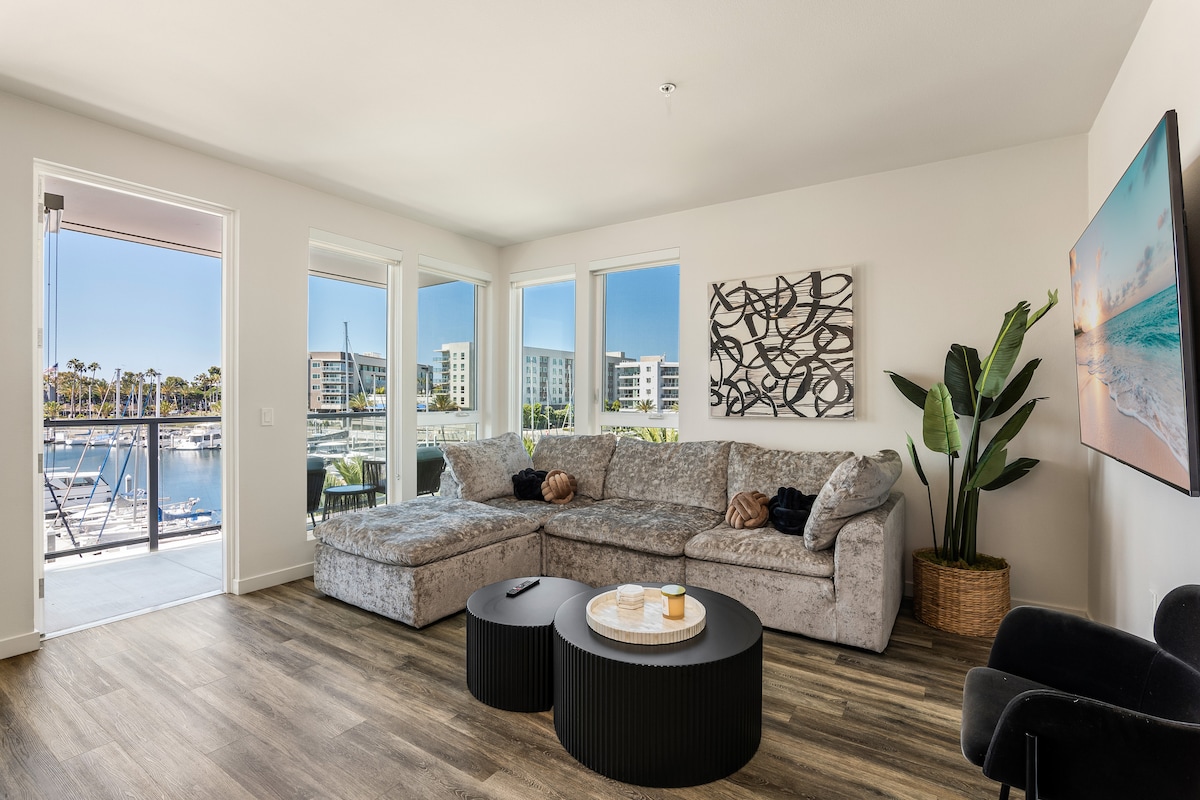 A spacious living area features a large sectional sofa arranged around two circular tables. Expansive windows provide views of the marina, allowing natural light to fill the room. Decorative art and a potted plant add a touch of elegance to the inviting space.