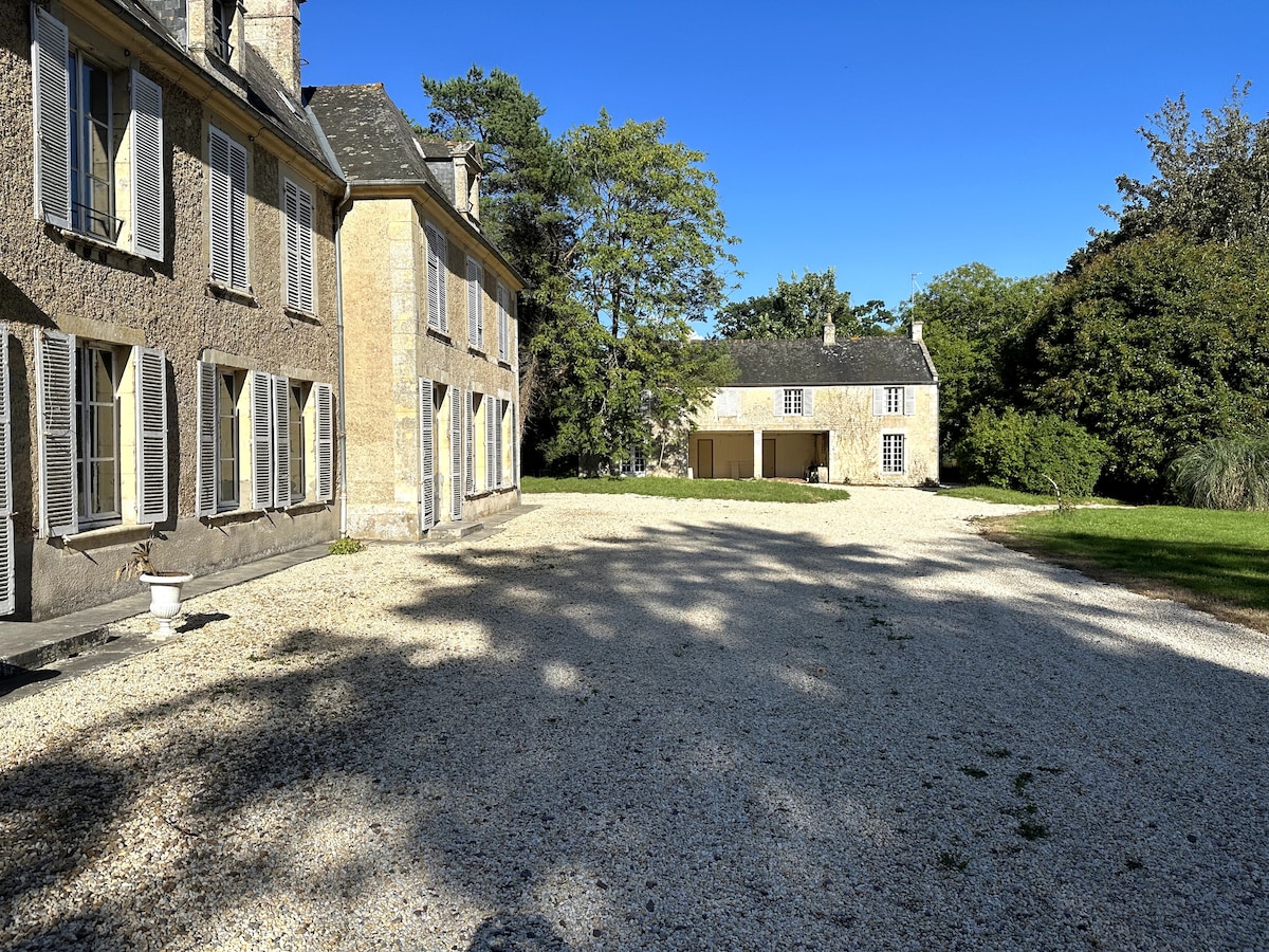 A gravel pathway leads to the historic stone house, framed by large windows that allow natural light to fill the interior spaces. A spacious lawn surrounds the home, with trees providing shade and a sense of tranquility.