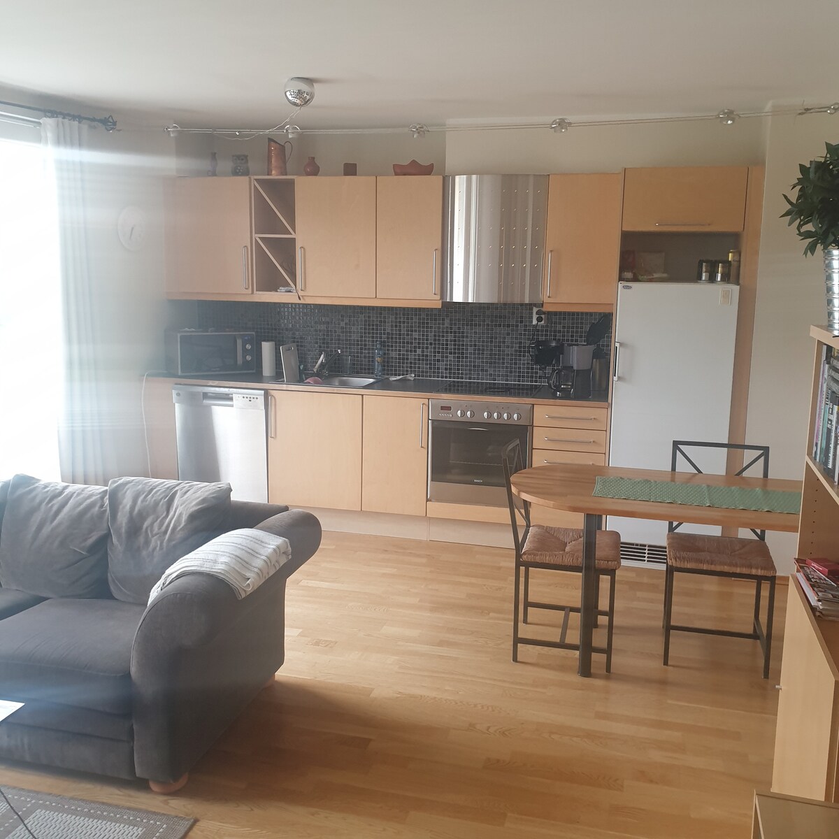 The modern kitchen area is equipped with wooden cabinetry and stainless steel appliances, including a refrigerator, oven, and dishwasher. A small dining table with two chairs is positioned beside a cozy couch. Natural light enters through large windows, illuminating the space.