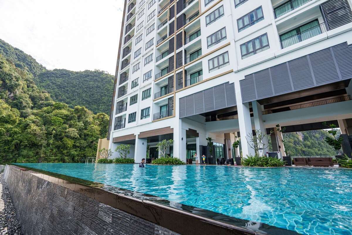 A modern building rises beside a serene infinity pool, reflecting the surrounding lush greenery. The pool's clear waters create a calming atmosphere, and seating areas are positioned nearby, providing space for relaxation and enjoyment of the natural surroundings.