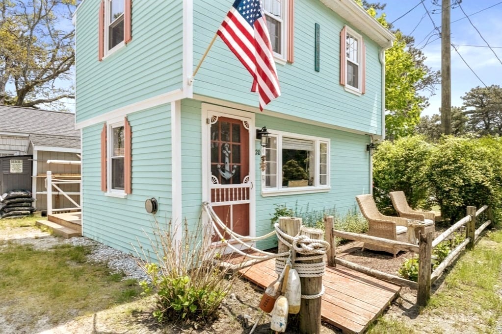 The charming exterior of a pastel blue cottage is showcased, featuring an American flag mounted above the entrance. Two wicker chairs are positioned on a small wooden deck surrounded by greenery, creating an inviting outdoor space. A wooden fence with decorative rope adds to the coastal aesthetic.