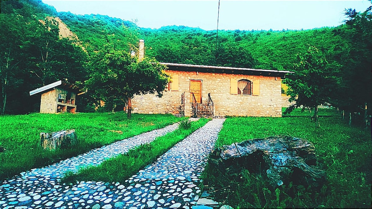 A charming stone house is set against a backdrop of lush green hills. A gently winding pebble pathway leads to the entrance, bordered by well-kept grass and natural landscaping. Nearby, a smaller building can be seen nestled among the trees.