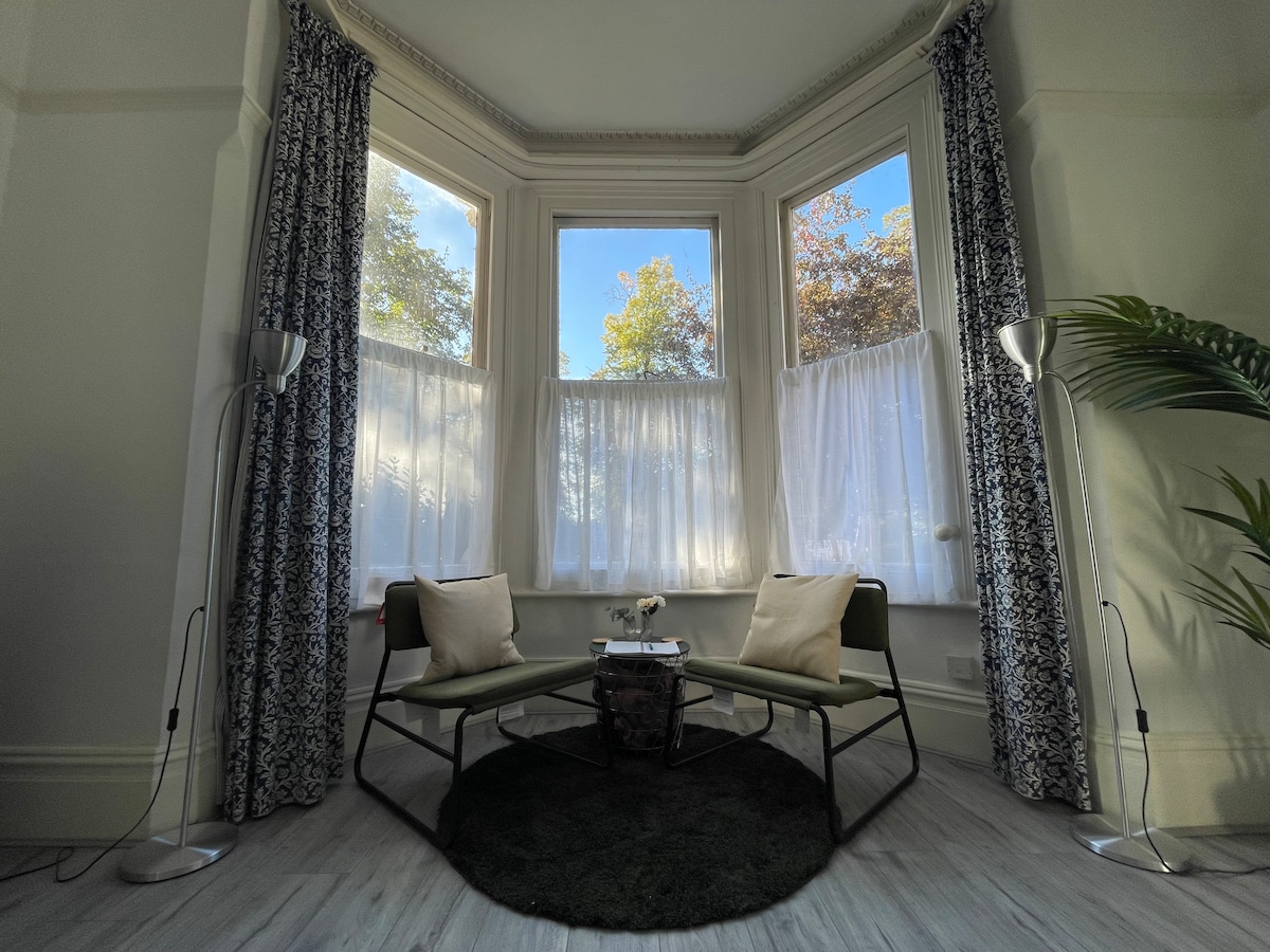 A bright corner of the living space features large bay windows, allowing natural light to fill the room. Two green chairs are placed beside a small table on a circular black rug. Sheer white curtains frame the windows, and a decorative plant adds a touch of greenery.