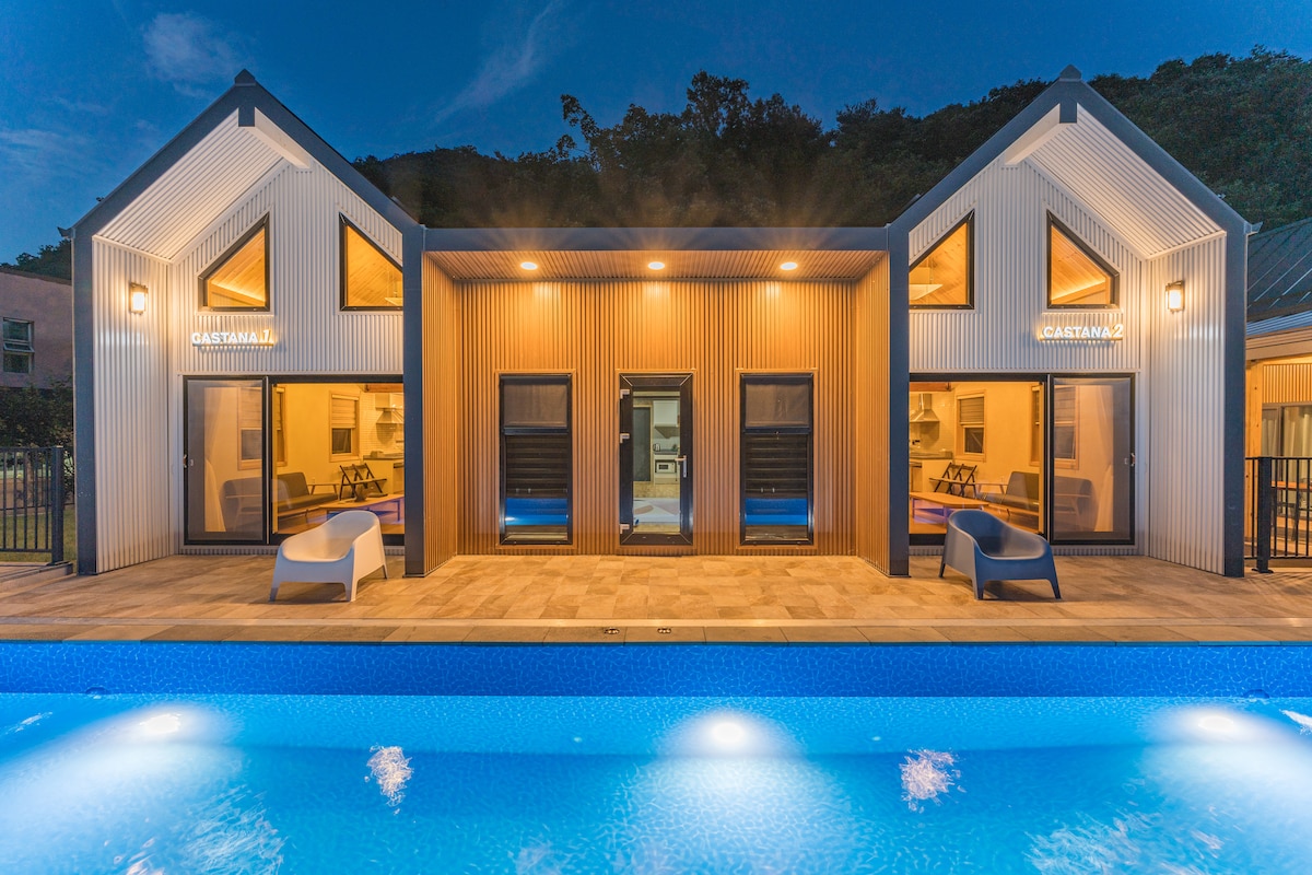 A two-story structure features large windows and inviting seating areas, set against a serene backdrop. The swimming pool is visible in the foreground, with ambient lighting reflecting off the water's surface, creating a calm atmosphere.