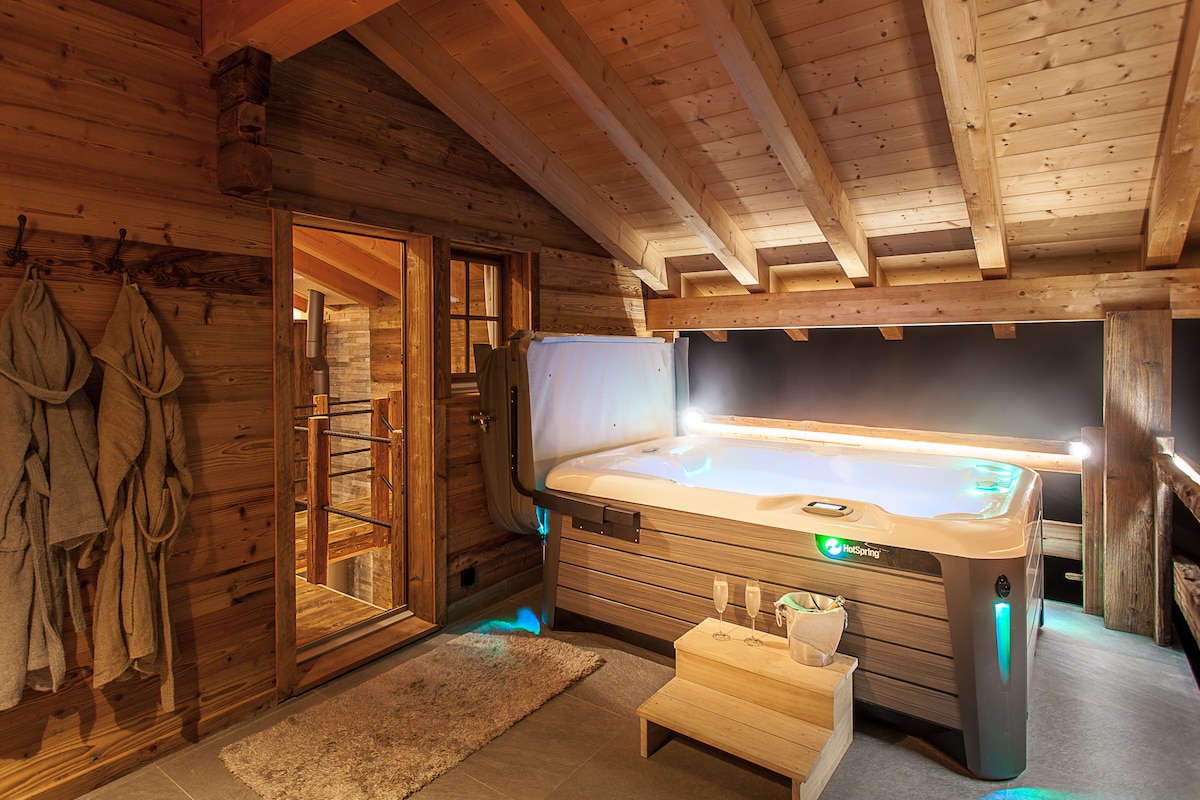 A private wellness area is featured, showcasing an outdoor jacuzzi with ambient lighting. The wooden ceiling beams add warmth to the room, which also includes natural stone flooring. A wooden bench and a soft rug complete the inviting space, enhancing relaxation.