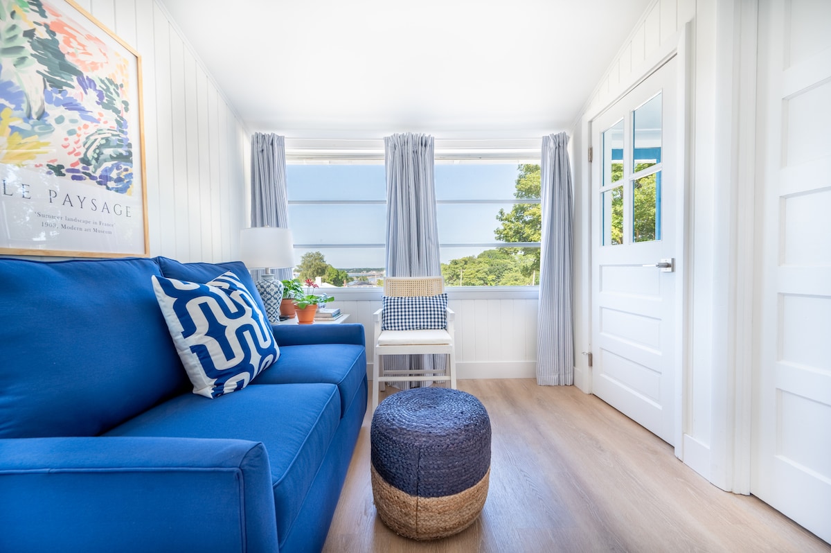 A bright and inviting sitting area features a blue sofa adorned with patterned cushions. A small white table with a plant stands nearby, and large windows allow natural light to illuminate the space, complemented by light-colored curtains.
