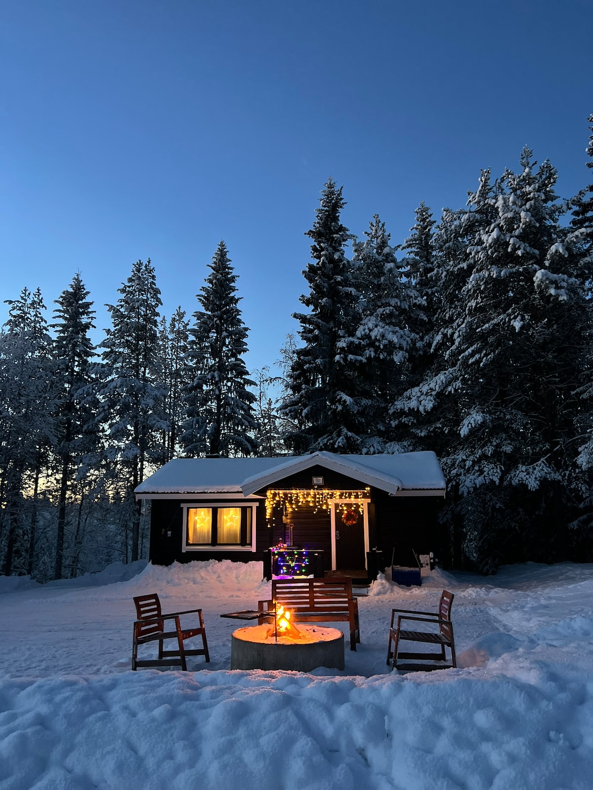 A cozy cabin is framed by tall, snow-covered trees, with a warm glow emanating from the window. Surrounding the fire pit, several wooden chairs are positioned for evening gatherings, enhancing the inviting winter atmosphere.