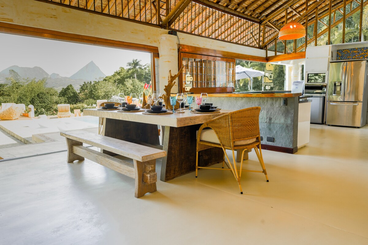 An open concept dining area features a large rustic wood table set with plates and glasses. Rattan chairs surround the table, and floor-to-ceiling windows provide a view of the surrounding landscape, including a prominent mountain in the background.