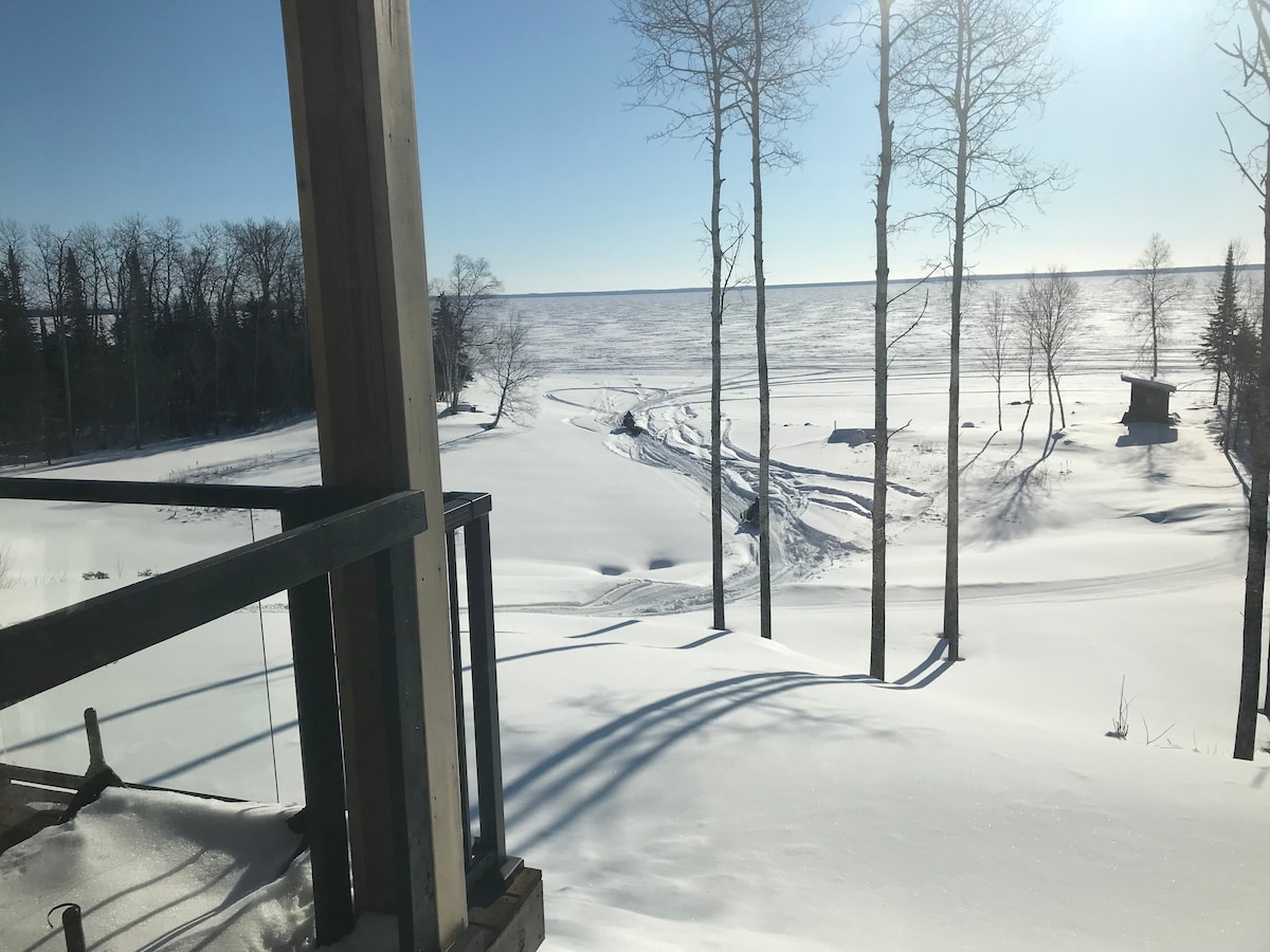 A serene winter landscape features a blanket of snow covering the ground, with the calm expanse of the frozen lake in the background. Trees stand tall on the horizon, casting soft shadows over the snow. A small structure is visible in the distance.
