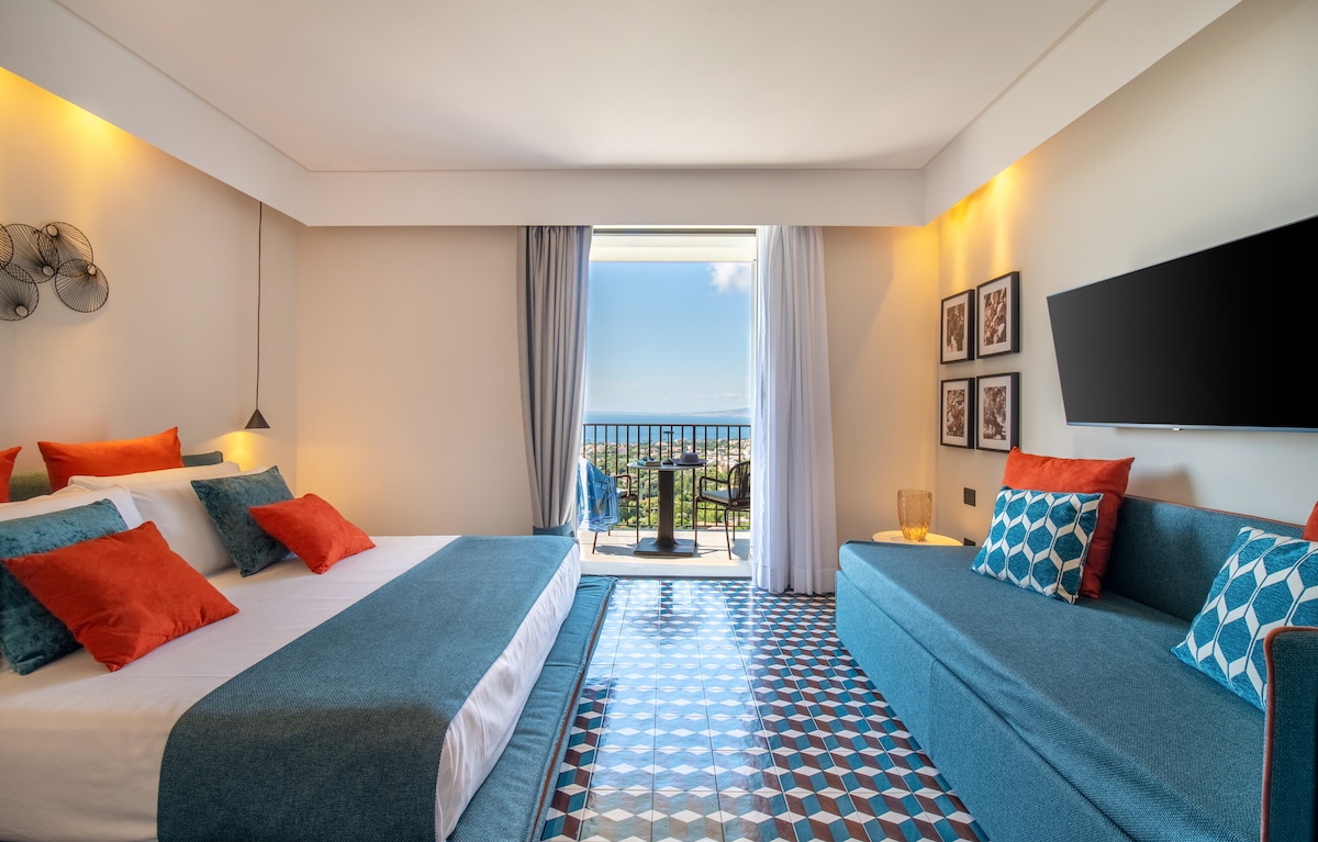 A spacious room features a large bed adorned with colorful pillows and a modern sofa. The tile floor reflects a geometric pattern, while a balcony with chairs offers a sea view. Light enters through large glass doors, enhancing the inviting atmosphere.