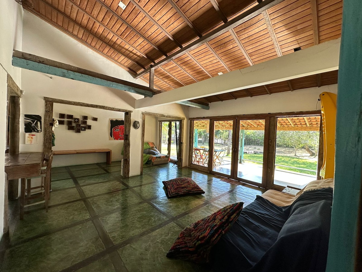 A spacious living area is characterized by high wooden ceilings and large glass doors, allowing natural light to fill the space. Colorful cushions are placed on the floor, and a wooden table is situated against the wall, creating a flexible and comfortable environment.