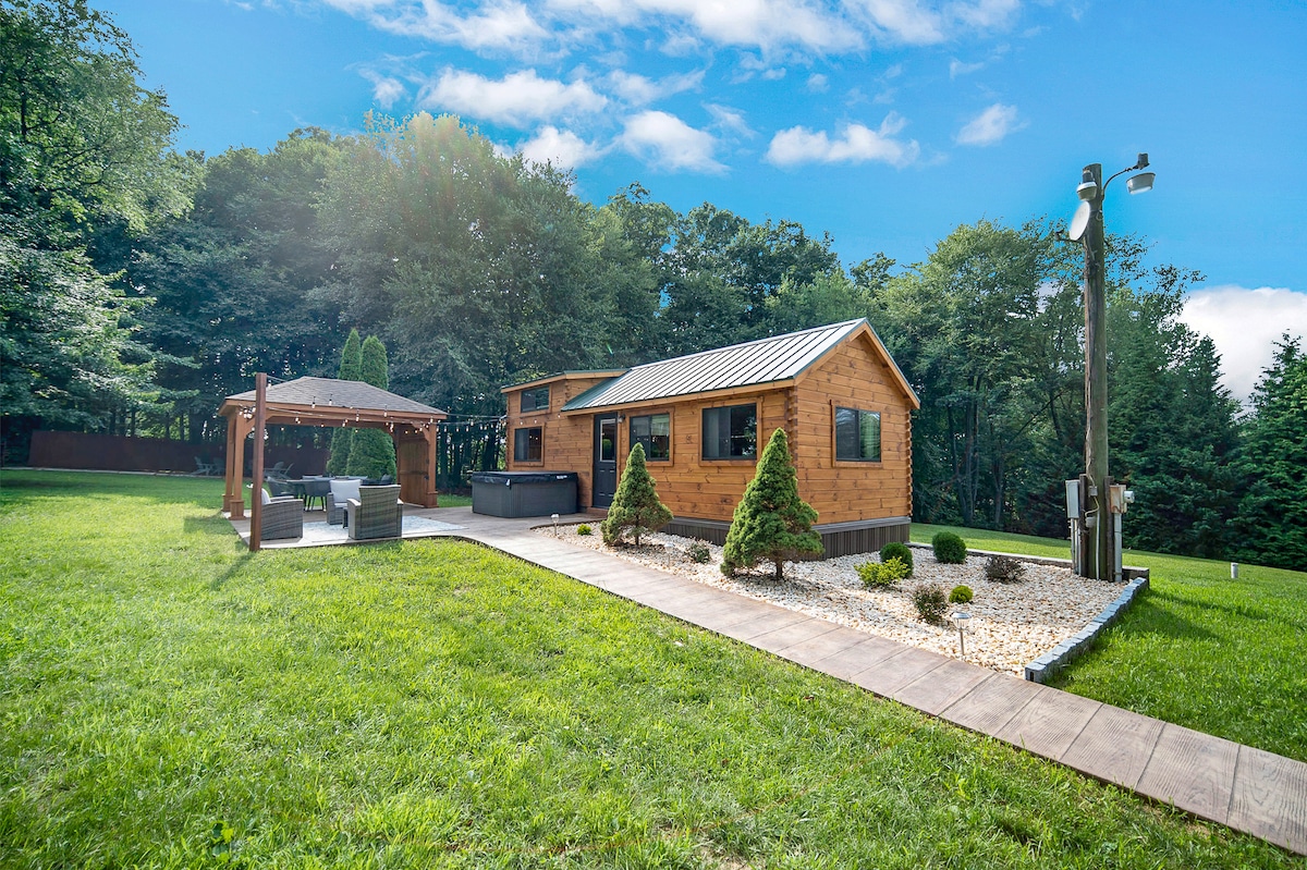 The exterior of a wooden cabin is surrounded by lush greenery, featuring a well-maintained lawn. A paved walkway leads to a spacious outdoor seating area under a gazebo, complemented by small hedges and landscaped stone arrangements.