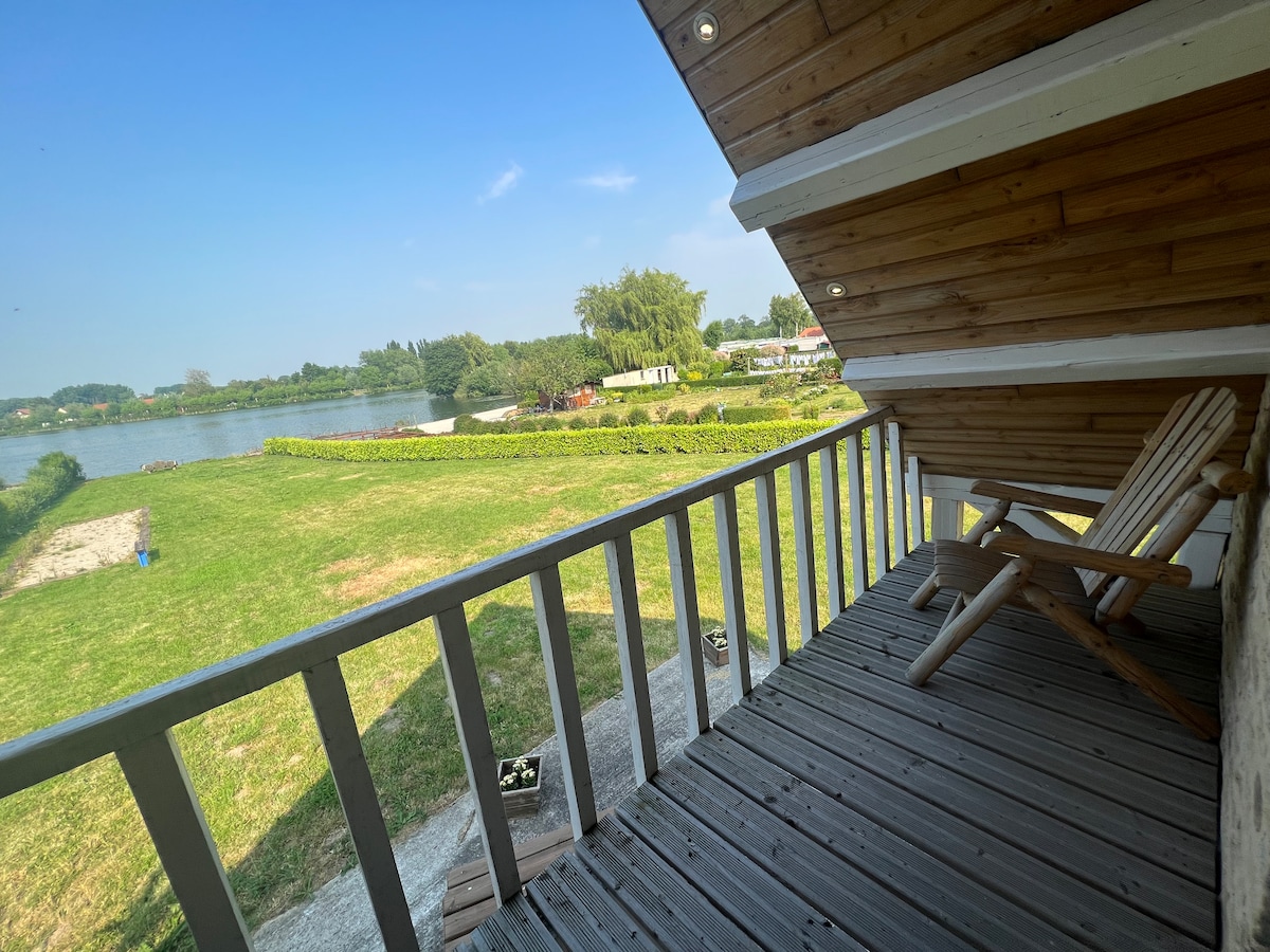 A wooden balcony offers a serene view of the surrounding grassy area and tranquil lake. Two deck chairs are positioned for relaxation, while a clear blue sky overhead enhances the peaceful atmosphere of the outdoor space.
