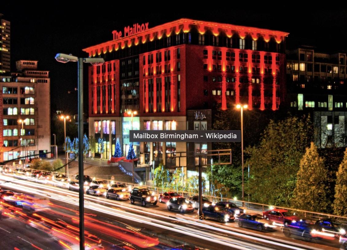 The Mailbox, a prominent building, is illuminated in red against the night sky. Traffic flows past with streaks of light from car headlights creating a dynamic scene. Decorative lights adorn the surrounding trees, adding to the urban atmosphere.