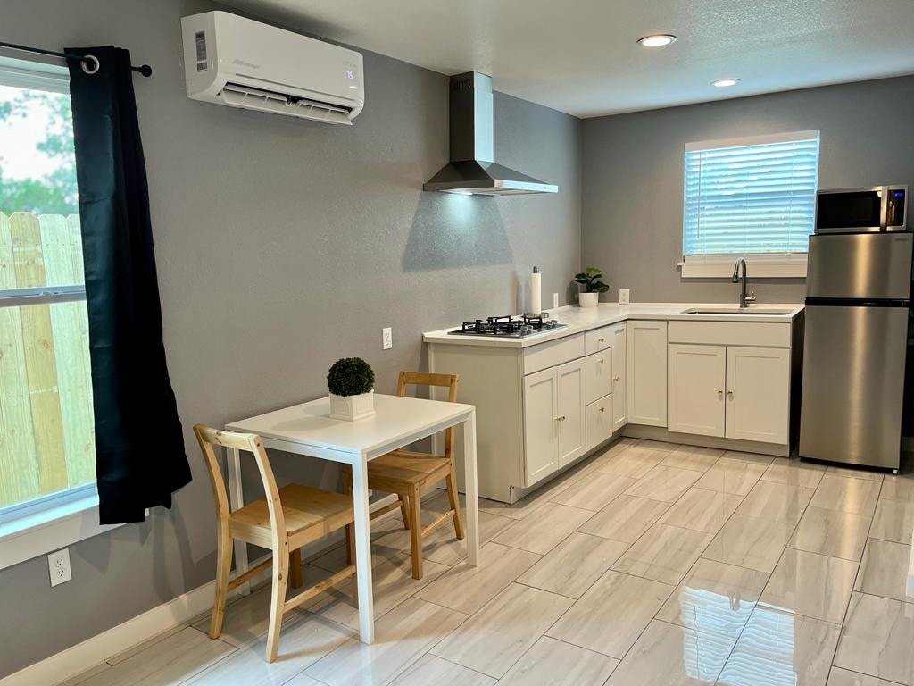 A modern kitchen space is displayed, featuring white cabinetry and a stainless steel refrigerator. A small dining table with two wooden chairs is positioned near a window, allowing natural light to brighten the room. The tiled floor reflects light, adding to the space's clean appearance.