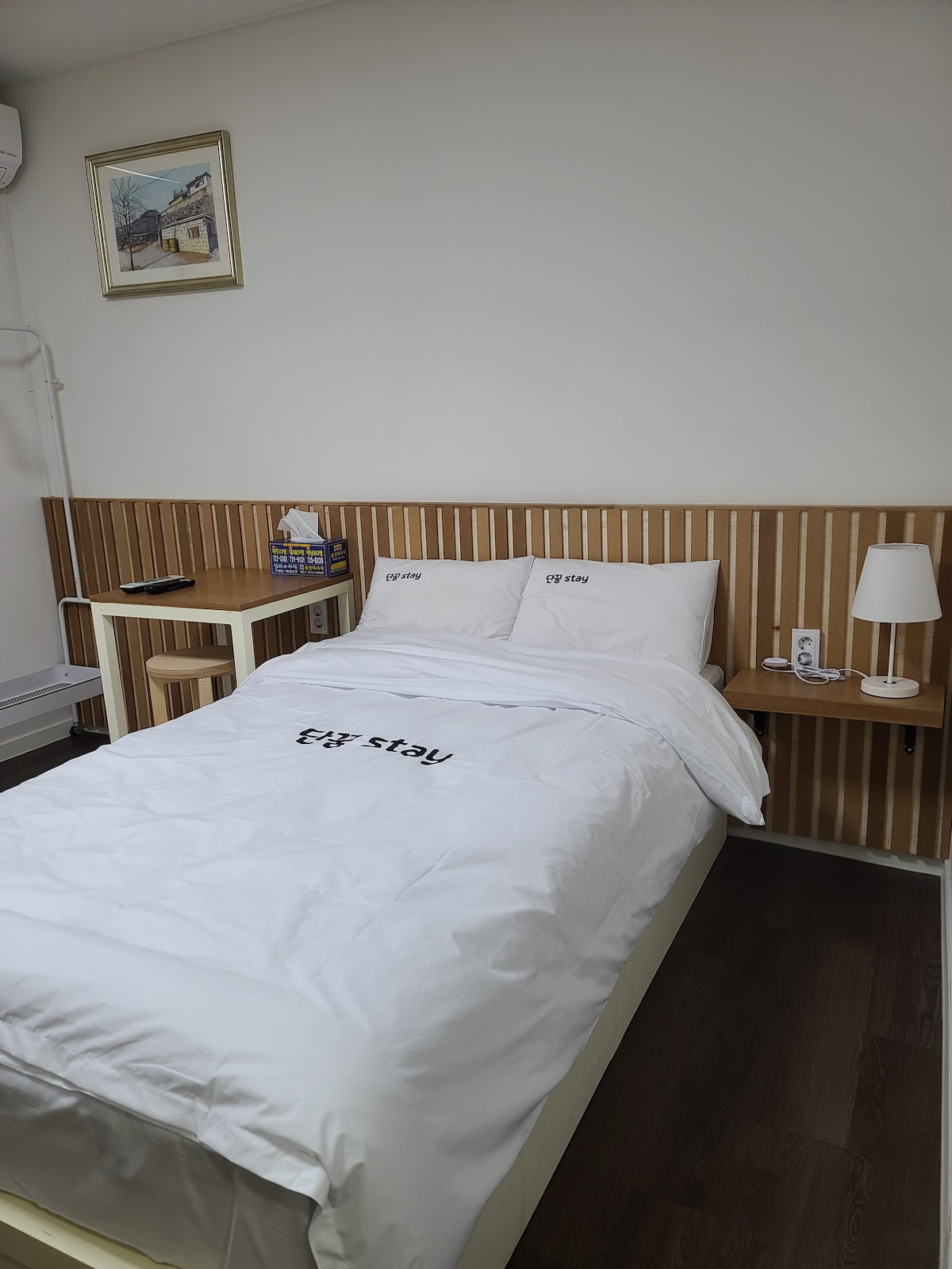 A compact guest room features a snug bed covered in a simple white blanket with a printed logo. A small wooden table and stool are positioned beside the bed. A wall lamp and an artistic wall hanging enhance the space, creating a modern and functional atmosphere.