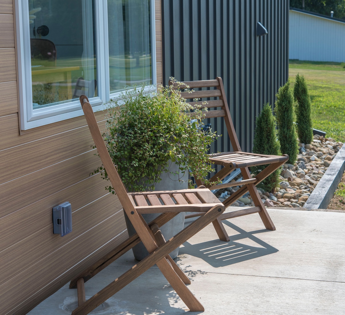 Two wooden folding chairs are positioned beside a flower pot, contributing to a relaxing outdoor space. A stone pathway leads to the house, with a neatly trimmed hedge visible in the background, enhancing the inviting entrance.