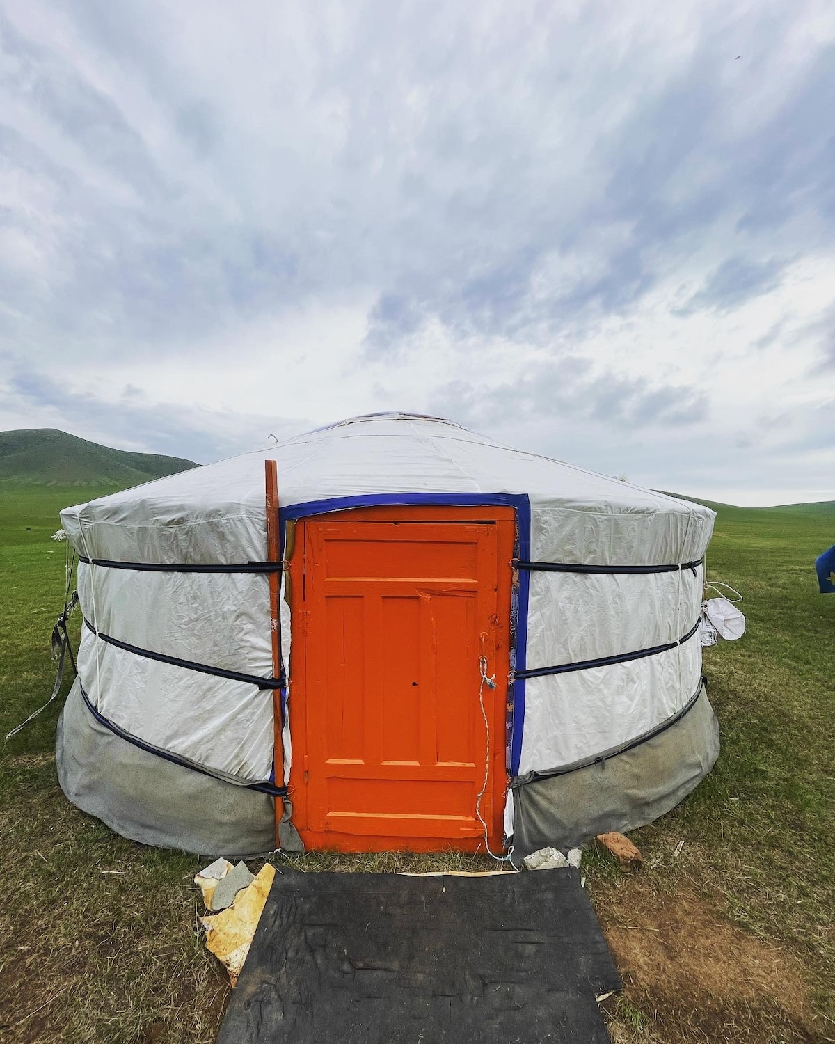 A round yurt is presented against a cloudy sky, featuring a distinct orange door that stands out against the white fabric. The structure sits on green grass, with the surrounding landscape gently rolling in the background.