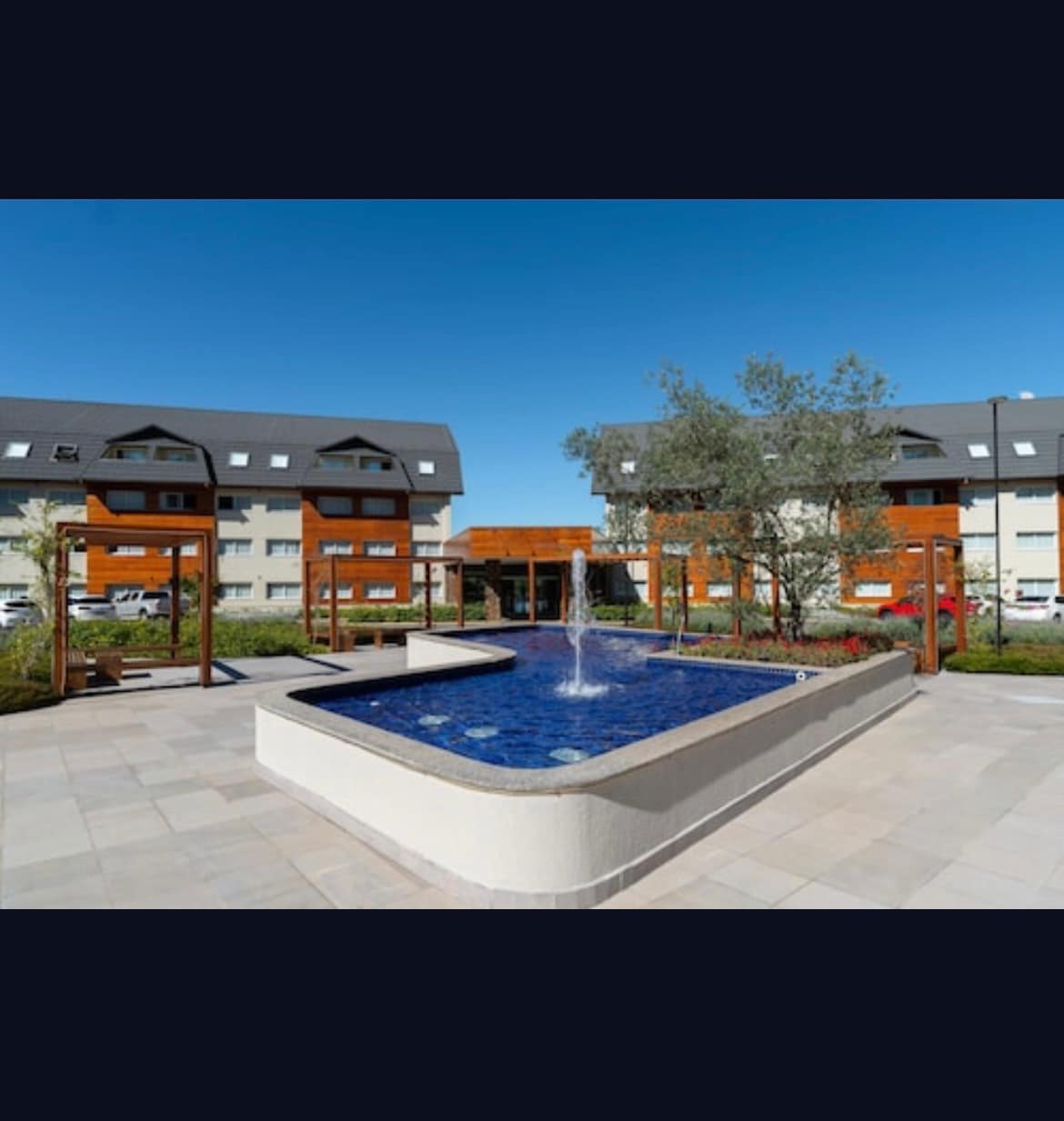 An outdoor area features a decorative fountain at the center of a rectangular pool, surrounded by landscaped greenery and pathways. The modern resort buildings provide a backdrop with large windows, enhancing the overall ambience of the space.