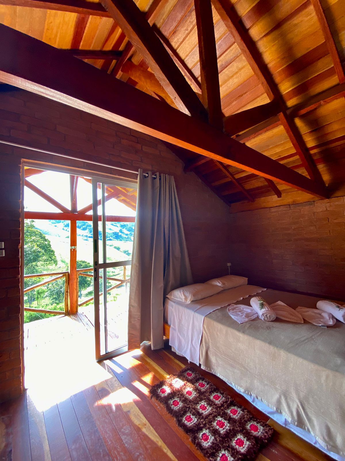 A bright sleeping area features a single bed with fresh linens and soft towels neatly arranged. Large glass doors open to a balcony, providing scenic views of the surrounding mountains. Exposed wooden beams enhance the rustic charm of the room.