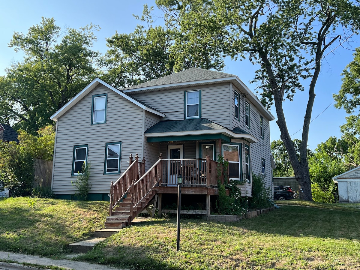 A two-story, light beige house is presented from its front angle, featuring a wooden deck with steps leading to the entrance. Large windows highlight the structure, surrounded by grassy areas and trees in the vicinity.