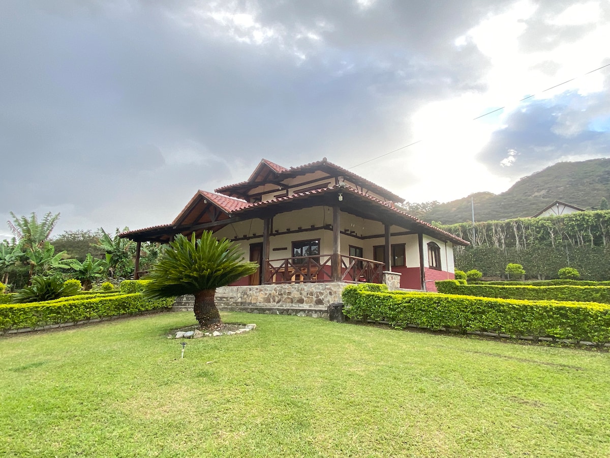 The house features a traditional architectural style with a red tiled roof and a spacious front porch. Lush green landscaping surrounds the property, complemented by neatly trimmed hedges and a palm tree in the foreground. Mountainous scenery forms the backdrop.