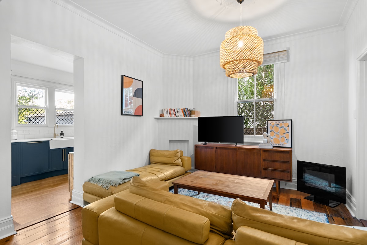 A spacious living room features a leather sofa and a wooden coffee table, with large windows allowing natural light to fill the space. A sleek television is positioned against a stylish wooden unit, complemented by a cozy fireplace and decorative wall art.