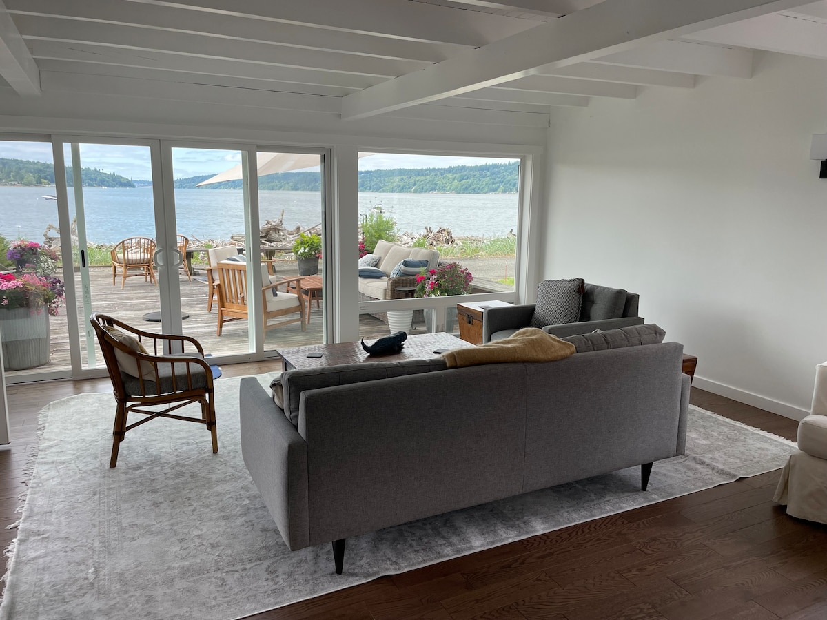 A bright living area features a comfortable gray sofa and a wooden armchair, with a soft area rug beneath. Large glass doors open to a waterfront patio filled with outdoor seating. The scenic view of the water and lush surroundings is visible through the doors.
