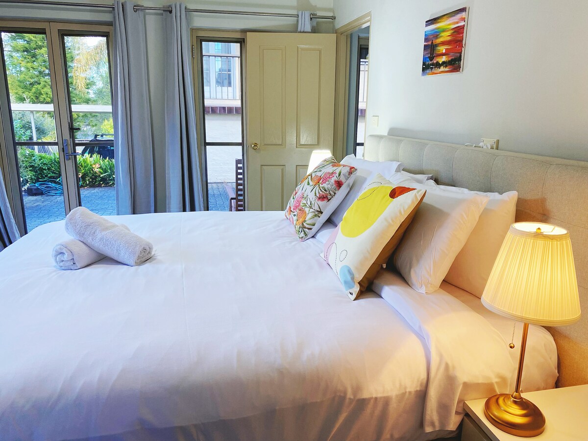 A spacious bedroom features a comfortable bed adorned with colorful pillows and a neatly rolled towel. A gentle light illuminates the room from a bedside lamp. Large windows allow for natural light and offer views of the outdoor garden.