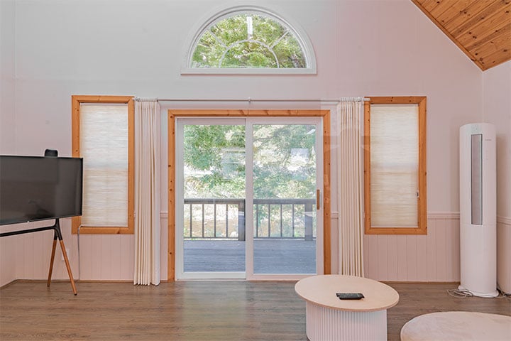 A bright living area features large sliding glass doors that open to a wooden deck. Natural light fills the space, highlighting the wooden beams of the ceiling and the white walls. A sleek entertainment unit, circular coffee table, and air conditioner complete the inviting setup.