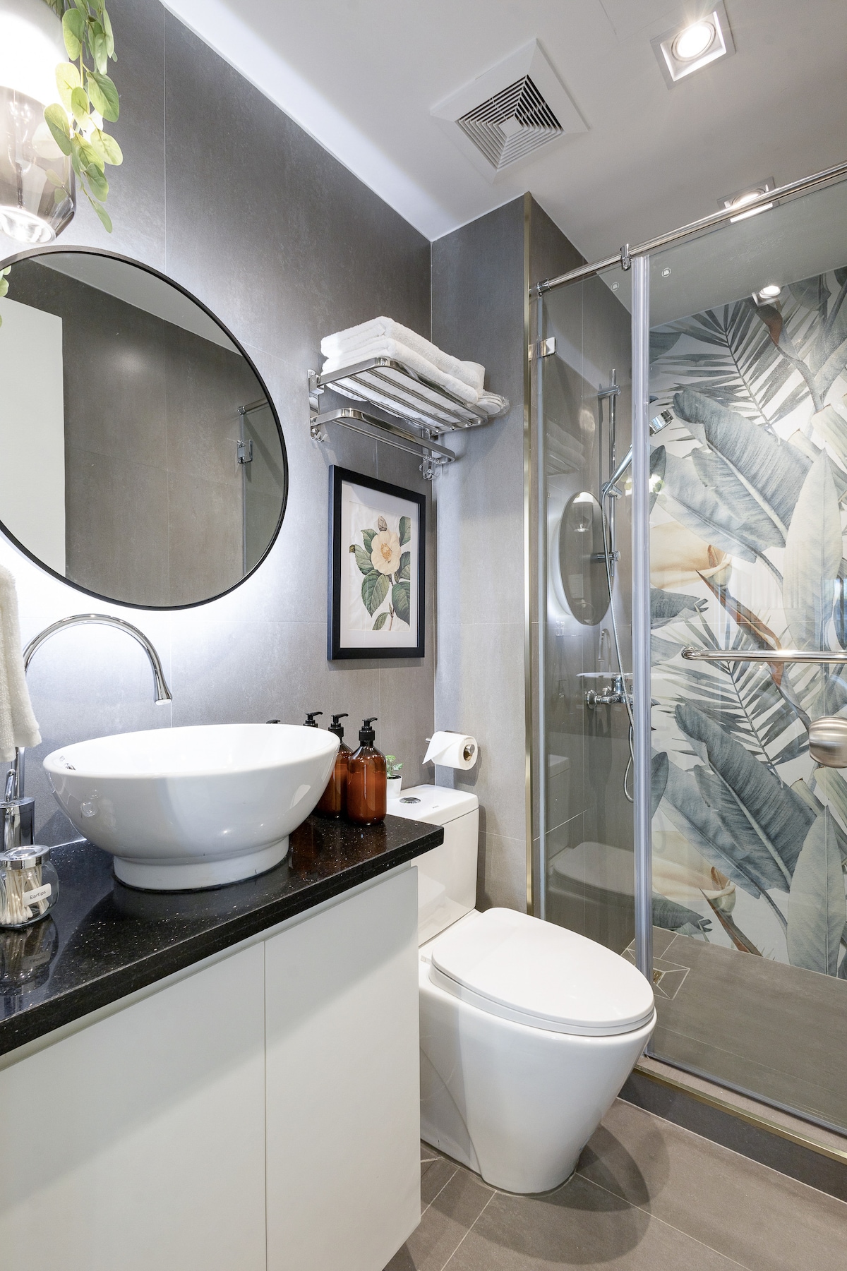 A modern bathroom features a sleek design with a circular mirror above a black countertop. A stylish vessel sink is paired with dark-colored soap dispensers. The shower area showcases tropical-themed tile art. Soft towels are neatly arranged on a shelf, adding functionality.