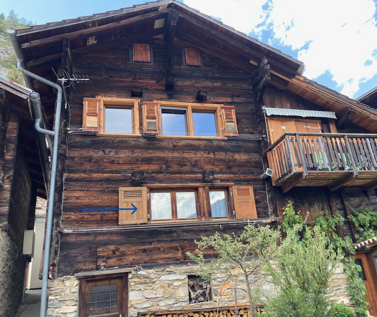 A charming wooden chalet is showcased, featuring multiple windows with traditional wooden shutters. A small balcony with a wooden railing is visible on the upper level. The façade is adorned with rustic stone at the base, surrounded by greenery, and a clear sky is seen above.