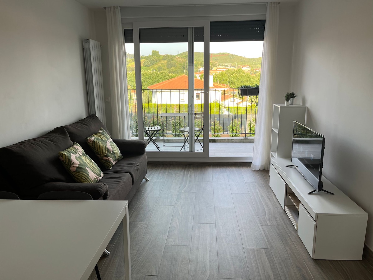 A comfortable living area is presented with a dark sofa adorned with patterned cushions. Large glass doors offer views of the surrounding landscape and provide access to a small balcony with chairs. A minimalist white entertainment unit sits across from the sofa.