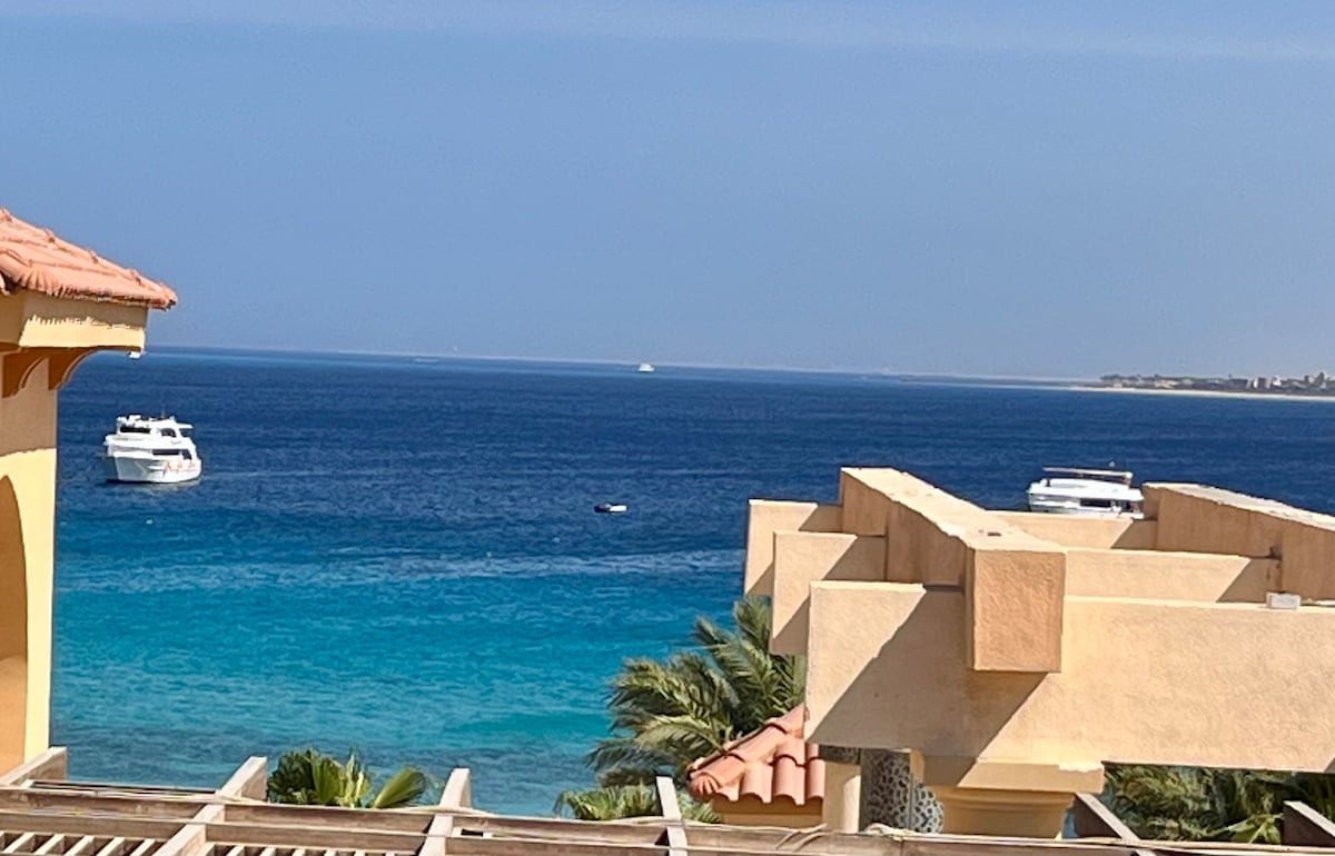 The image presents a serene view of the turquoise sea, with several boats anchored in the distance. Lush palm trees are visible in the foreground, providing a tropical ambiance, while the bright blue sky complements the tranquil waterscape.