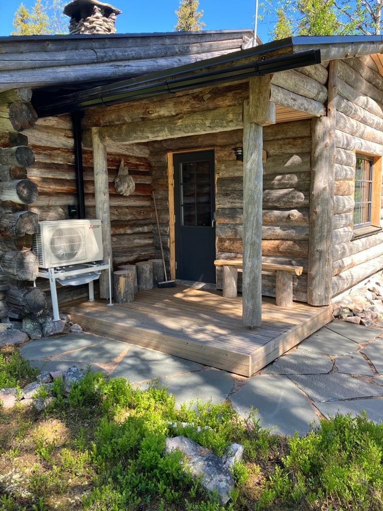 The cabin entrance features a wooden porch with log accents and a dark door. Stone slabs create a path leading up to the entrance, and an air conditioning unit is situated beside the steps for convenience. Surrounding greenery adds a natural touch.