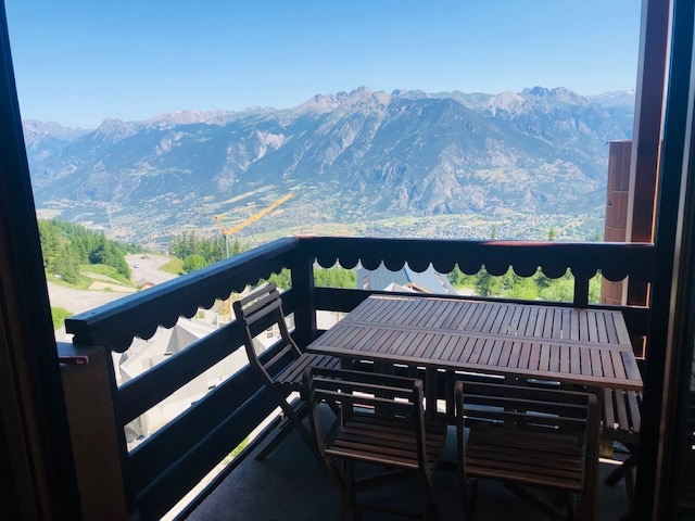 A balcony features a wooden table surrounded by several folding chairs, with expansive views of the valley and mountains in the background. Clear blue skies enhance the scenic landscape, providing an inviting outdoor space for relaxation.