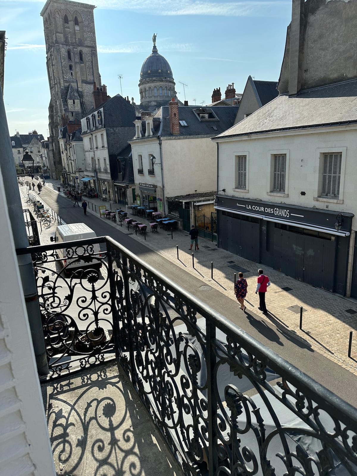 A wrought iron balcony railing frames a view of a bustling street lined with historic buildings. The Basilica of St. Martin is visible in the distance, with its towering bell tower and dome. Pedestrians stroll along the cobblestone street, showcasing the vibrant atmosphere.