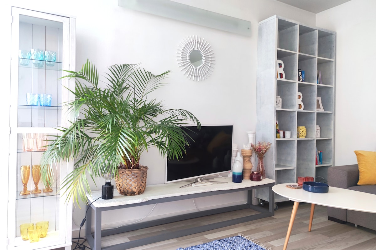 A modern living area features a television mounted on a light-colored wall. A tall green plant adds a natural touch beside a console table. Decorative items, including vases and glasses, are arranged on shelves and in a display cabinet, creating a tidy and welcoming environment.