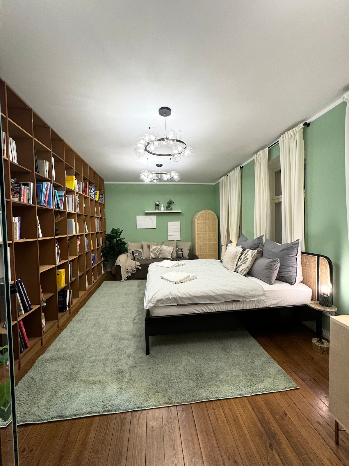 A serene bedroom features a spacious bed adorned with various decorative pillows. A bookshelf spans one wall, filled with books and decorative items. Light oak flooring complements a soft green area rug, while curtains frame large windows, allowing for natural light to enter.