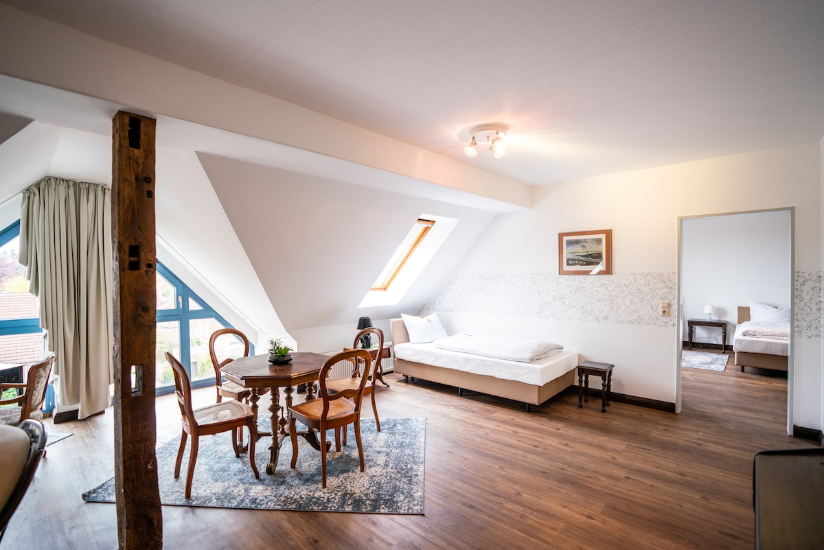A spacious attic room features natural light from skylights and large windows. A dining table surrounded by four chairs is positioned near a decorative area rug. A single bed sits against the wall, with a light-colored sofa in the corner.