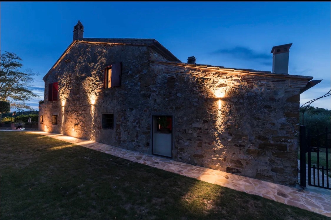 An exterior view of a rustic stone building is illuminated at night. Warm lights highlight the textured stone walls and features, creating a soft ambiance. A pathway leads to the entrance, with a grassy area surrounding the structure, enhancing the tranquility of the setting.