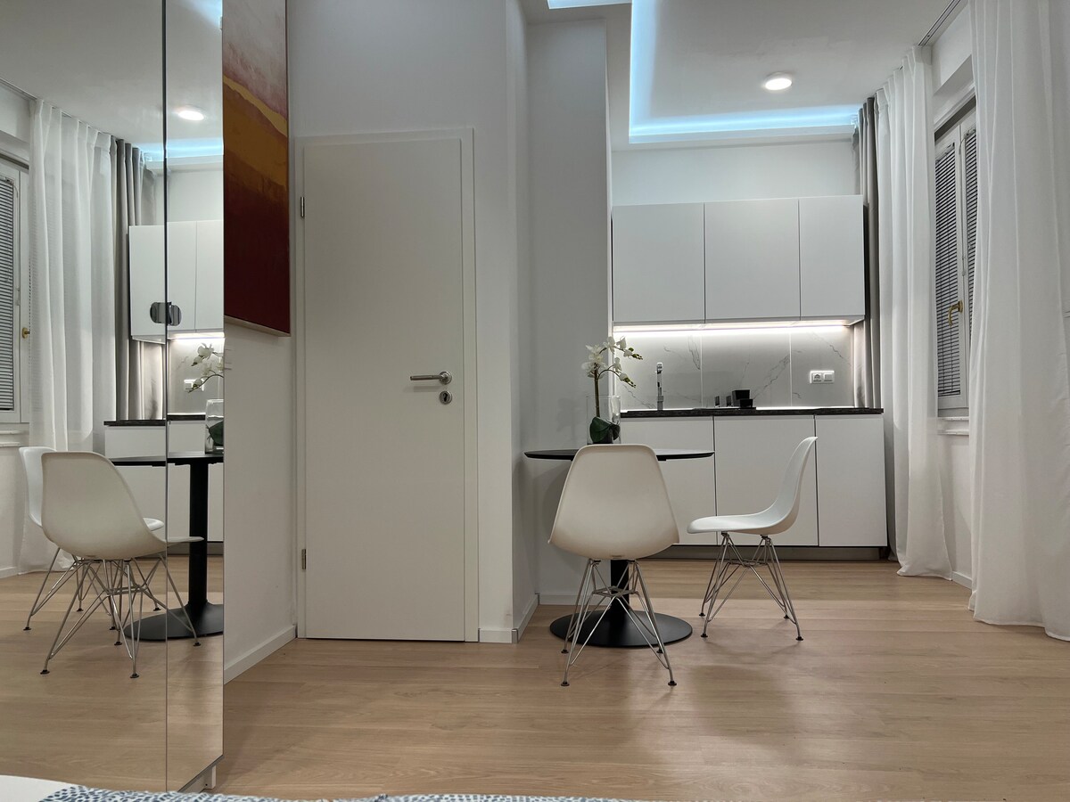 The studio features a compact dining area with two white chairs around a circular table. Modern cabinetry is visible in the background, accompanied by bright overhead lighting. Natural light enters through a window, and soft, neutral tones are present throughout the space.