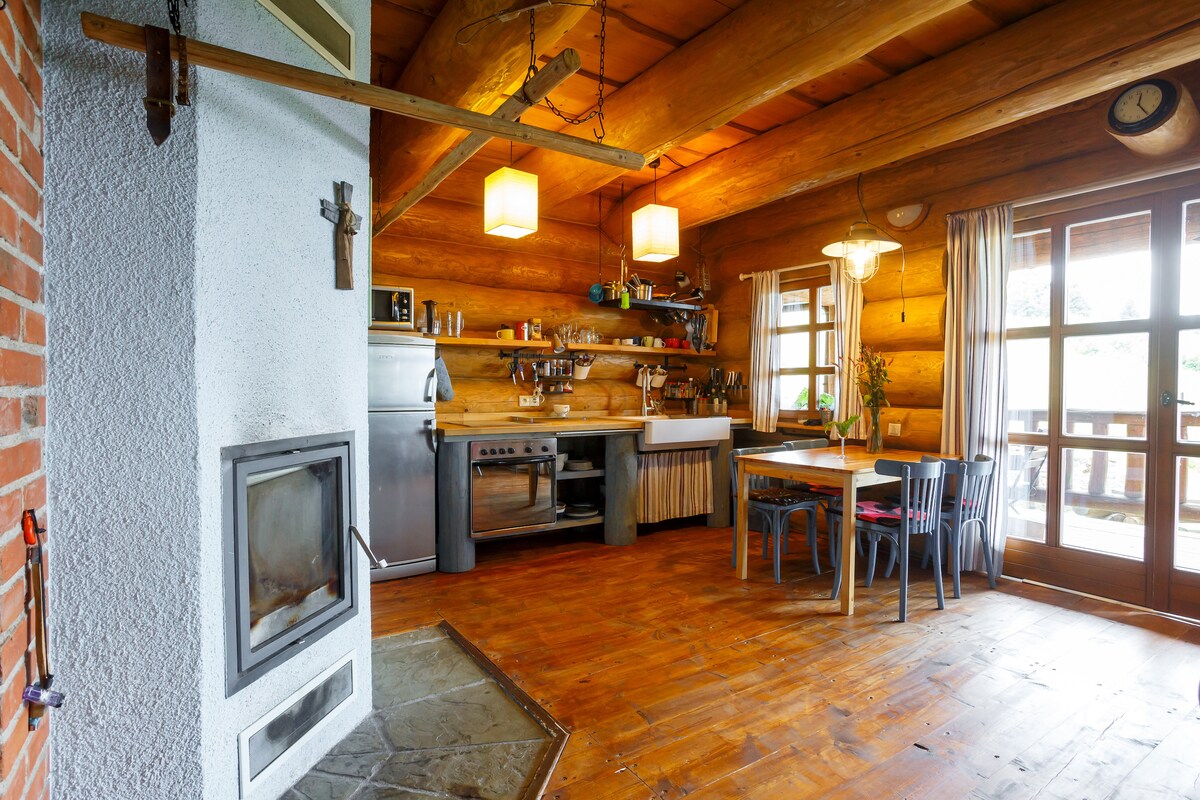 A rustic kitchen is showcased with wooden beams and a stone fireplace. Metal appliances line the walls, complementing a wooden dining table surrounded by chairs. Natural light filters through the glass doors, leading to a terrace, creating an inviting space for meal preparation and dining.