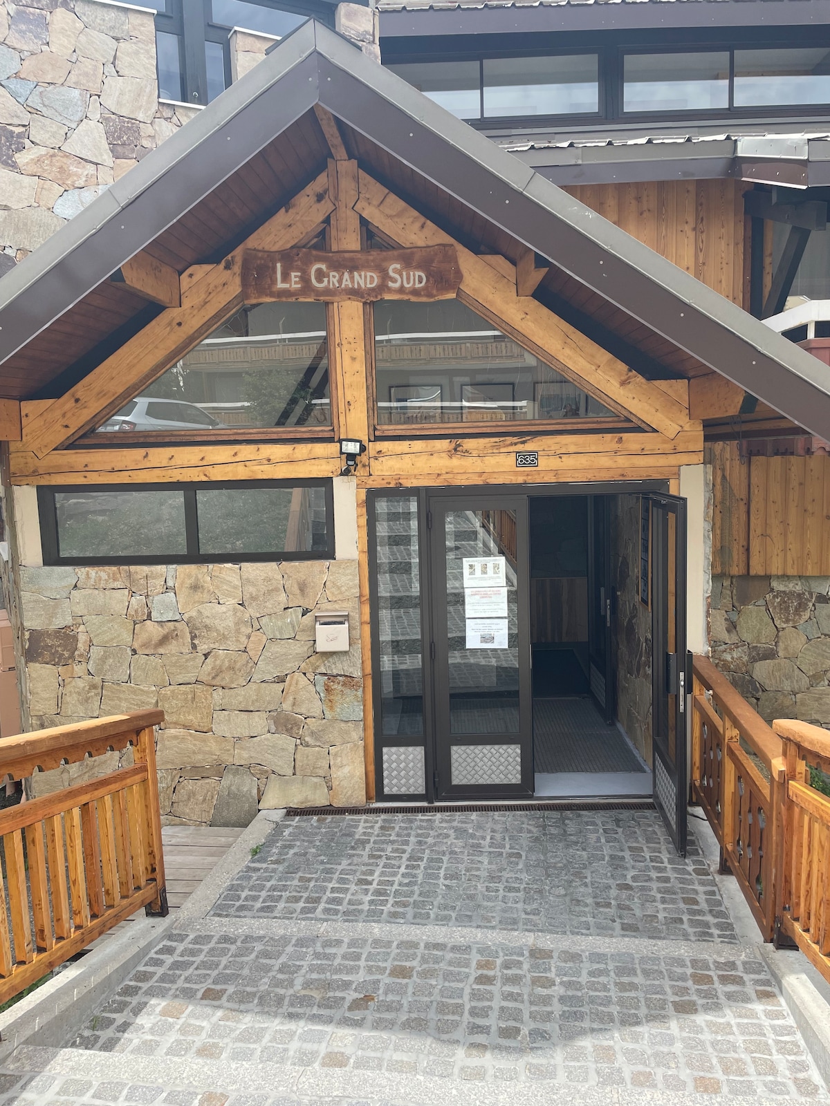 The entrance of 'Le Grand Sud' is framed by natural stone and wooden accents, creating an inviting facade. Double glass doors provide access, with a wooden sign displaying the name prominently above. A stone pathway leads up to the entrance, surrounded by rustic railings.