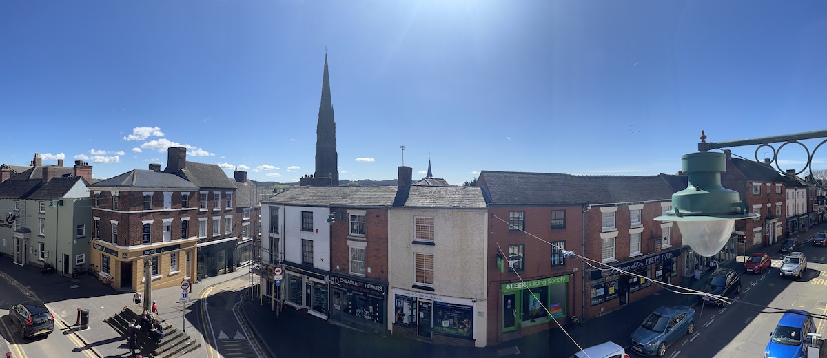 A panoramic view of the town center is captured, featuring historic buildings with a mix of brick and painted facades. In the distance, a prominent church steeple reaches towards the sky under bright sunlight, while cars are parked along the street.