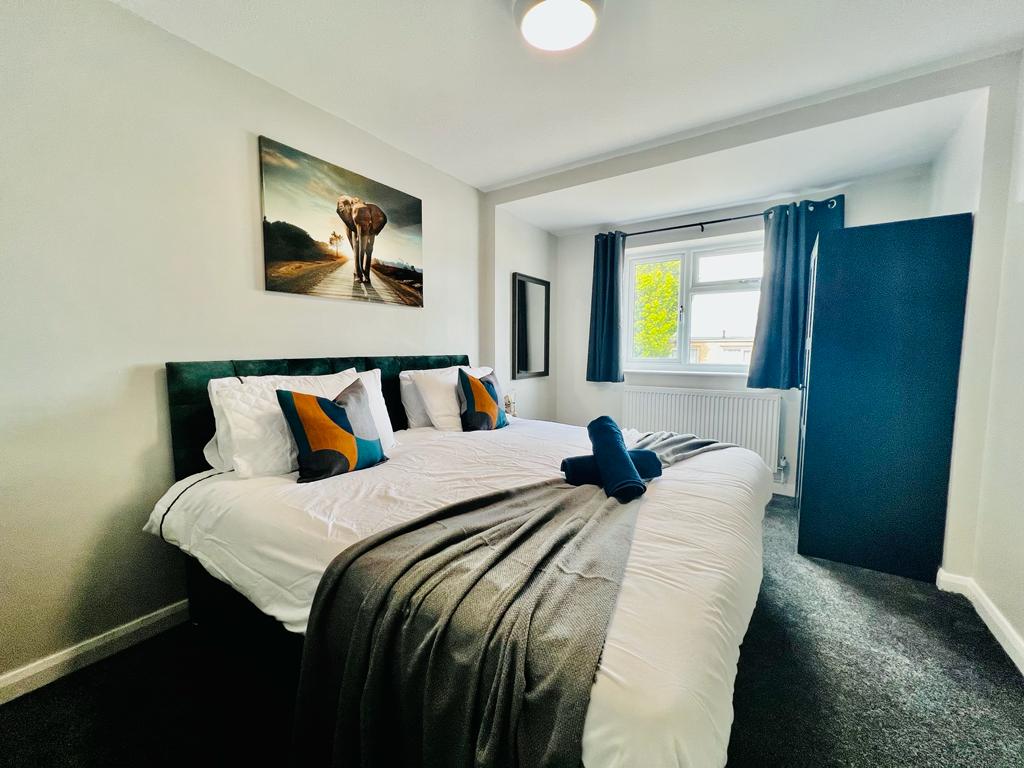 A spacious bedroom features a large bed adorned with colorful accent pillows and a cozy blanket. Natural light filters through a window, and an artwork of an elephant is displayed above the bed. A wardrobe is positioned nearby, enhancing the room's functionality.