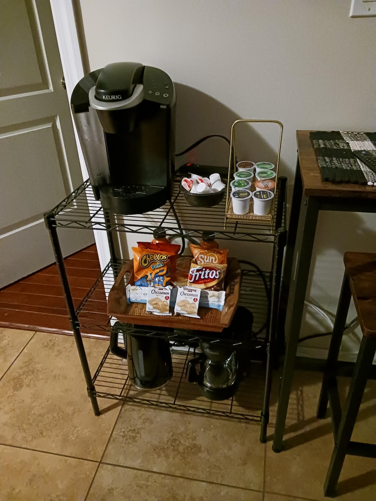 A coffee station is presented on a metal shelving unit, featuring a Keurig coffee maker, various coffee pods, and creamers. Snack items, including individual bags of chips, are arranged on a wooden tray, with two thermal carafes placed below for coffee storage.
