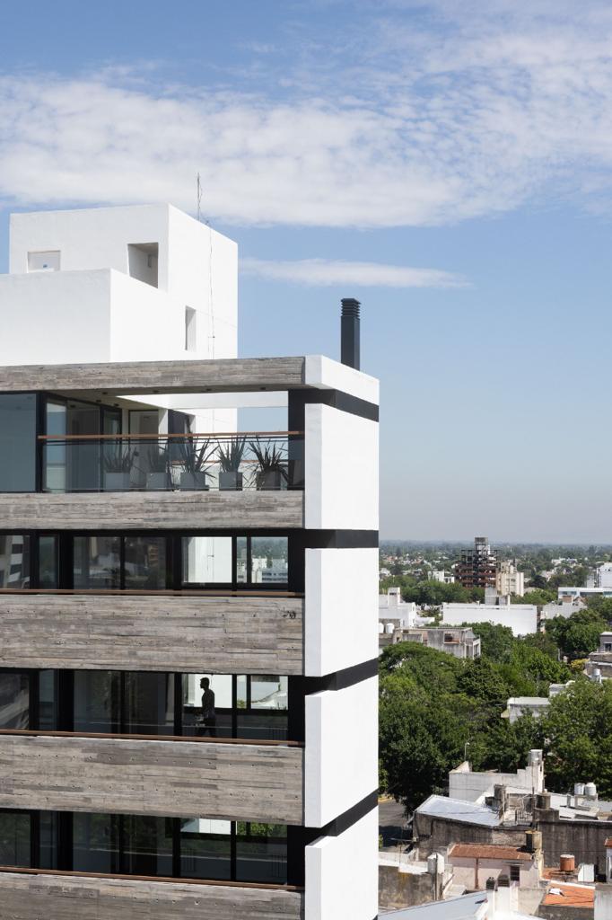 A modern building with a striking design features wooden elements and large glass windows. Plants are arranged on the terrace, enhancing the outdoor space. A clear sky and distant cityscape are visible in the background, providing a sense of openness and light.
