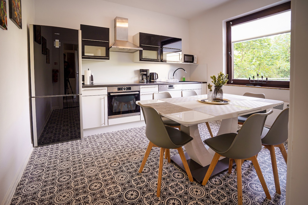 A modern kitchen is equipped with sleek black and white cabinetry, featuring a gas stove and stainless steel appliances. A dining table is surrounded by four grey chairs, positioned on patterned tile flooring. Natural light filters through a large window, illuminating the space.